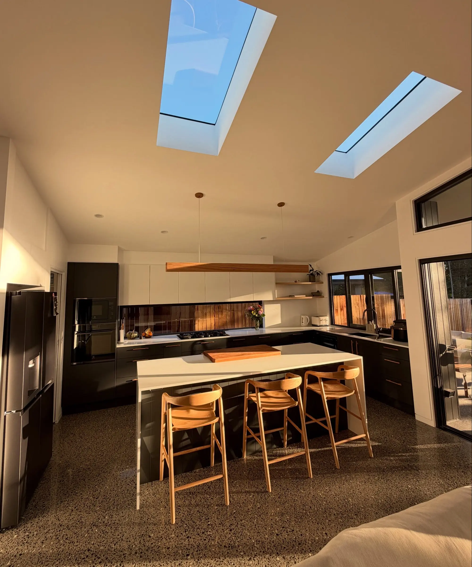 Kitchen in low golden sun: dark base cabinets and tall units, white overhead cupboards, a white stone island with a waterfall end and three timber stools, a long timber pendant, two skylights in a raked ceiling and a polished concrete floor.